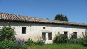 A stone building with a tiled roof, small windows, and a garden with various plants.