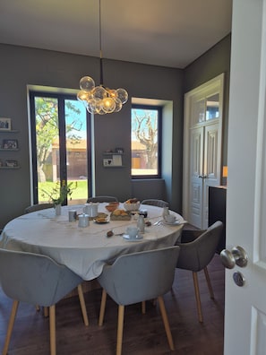 A dining area with a round table set for four, chairs, a chandelier, and a view of the outdoors through large windows.