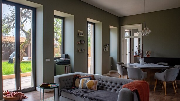 Una sala de estar moderna con un sofá gris, un comedor con una mesa redonda y sillas, y ventanas amplias que ofrecen vistas al exterior.