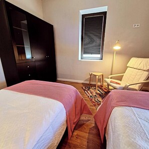 A bedroom with two beds, a window with blinds, a chair, and a lamp.