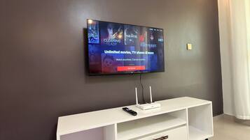 A flat-screen TV mounted on a dark wall, a white entertainment unit with a remote control, and a white curtain on the right.