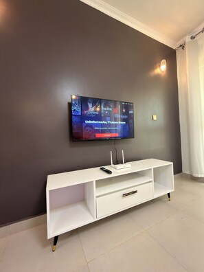 A flat-screen TV mounted on a dark wall, a white entertainment unit with a remote control, and a white curtain on the right.