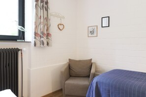 Ein Zimmer mit Heizkörper, einem Sessel, einem Bett mit gestreifter Bettdecke, einem Fenster mit Vorhängen und zwei gerahmten Bildern an der Wand.