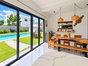 A modern interior with a large glass door leading to a pool, a wooden dining table, and a hanging light fixture. - 3 bedroom villa,5 minutes to the main beach driving 3 minute to other beach (Cabarete)