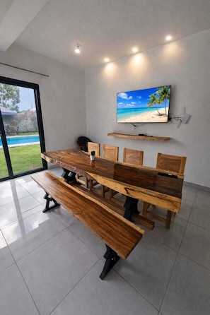 Ein moderner Essbereich mit Holztisch und Bänken, einem an der Wand montierten Fernseher und Blick auf einen Pool im Freien.