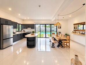 Et moderne køkken med en central ø, et spiseområde med et træbord og bænke, samt udsigt til en swimmingpool gennem glasdøre.