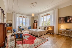 A bedroom with a bed, two chairs, a desk, a dresser, and a large window with curtains.