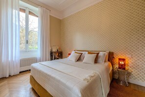 Quarto com cama grande, janela com cortinas, papel de parede estampado e mesa de cabeceira com abajur.