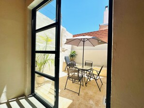 Blick durch ein Fenster auf eine Terrasse im Freien mit Tisch, Stühlen, einem Blumentopf und einem großen Sonnenschirm.