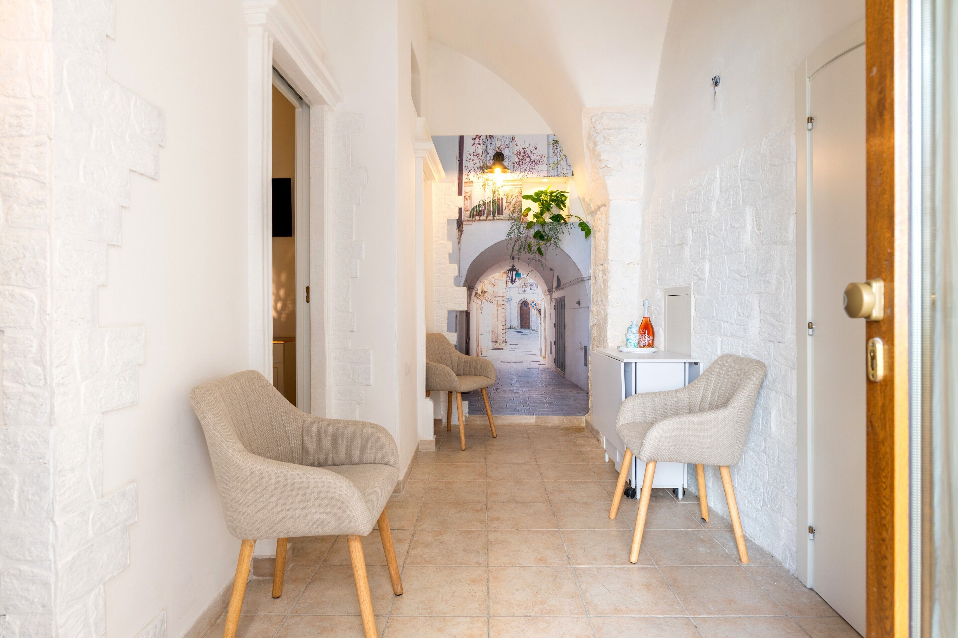 A hallway with white brick walls, beige chairs, and a view into a dining area with a table and chairs.