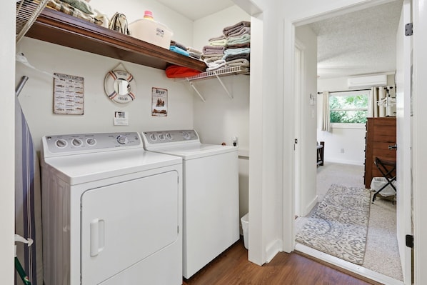 A laundry room with two white front-loading washers and dryers, a shelf above with folded towels, and a welcome sign.