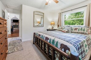 A bedroom with a bed, a nightstand, a lamp, a window with a view of trees, a mirror, and a wooden dresser.
