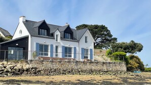 A coastal house with a stone wall foundation, multiple windows, and a prominent tree. - Waterfront 4* villa on the Etel ria. Ideal for families. (Plouhinec)