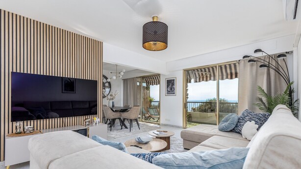 A modern living room with a flat-screen TV, a sofa, a dining area, and a balcony with curtains. - MC Riviera: Modern Apartment With Stunning sea View and Terrace Near Monaco (Beausoleil)