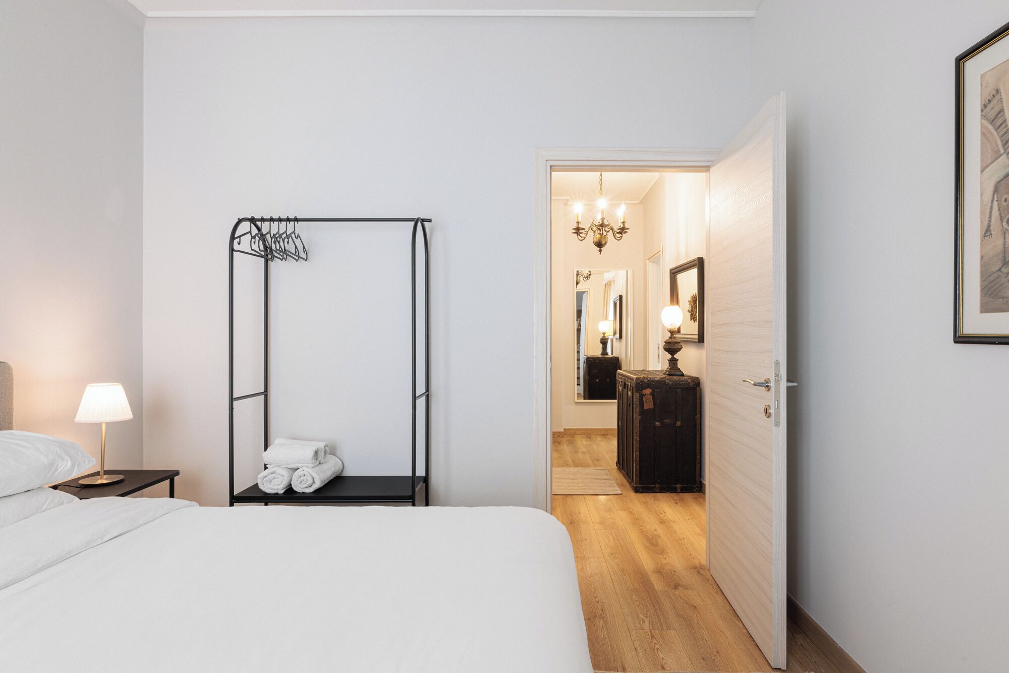 Ett hotellrum med en säng, ett metallställ med handdukar, ett nattduksbord, en lampa och en dörr som leder till en hall med en spegel och en byrå.