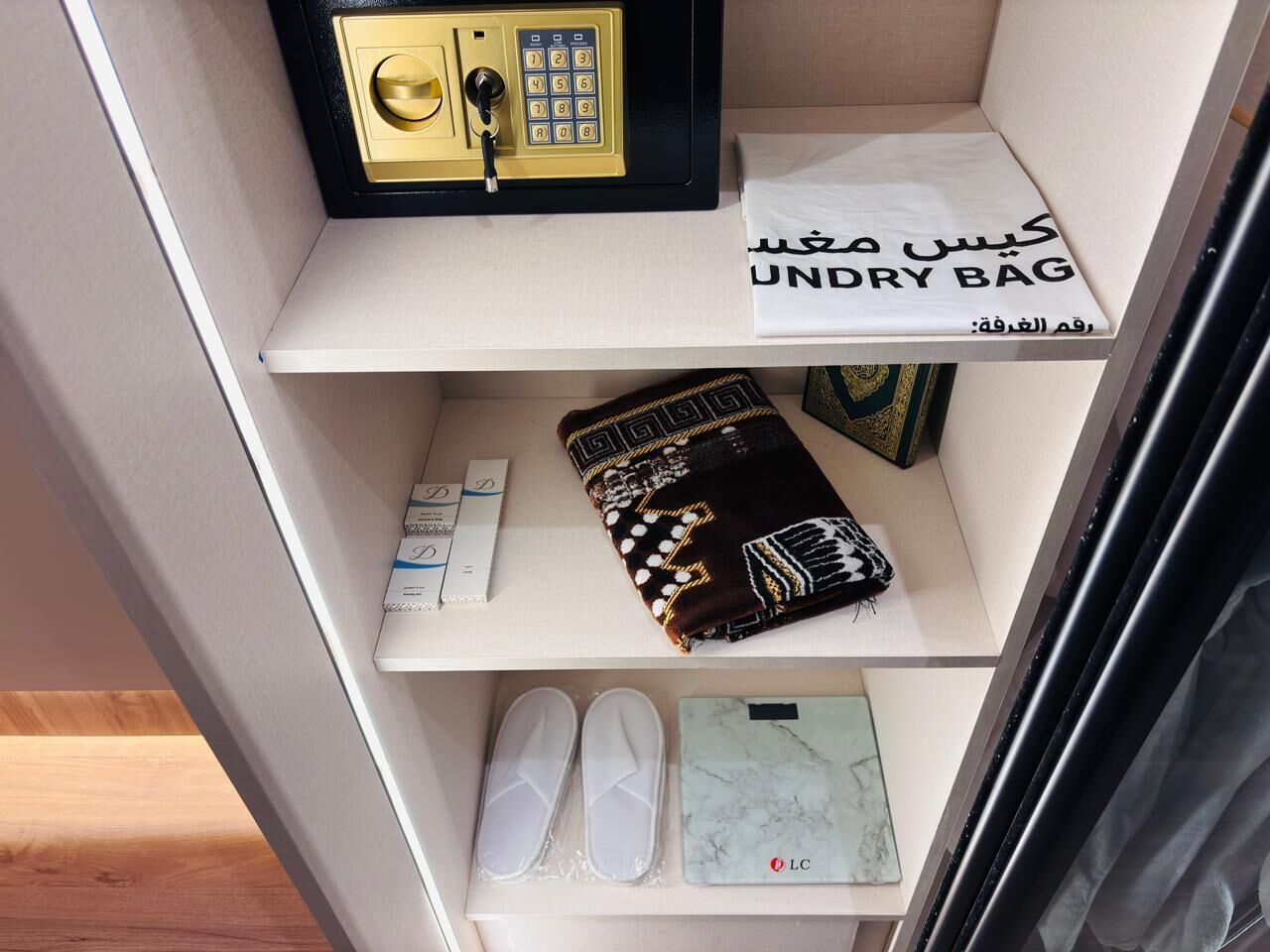 A shelf with a safe, a white bag with Arabic text, a patterned cloth, a box of tissues, a pair of white slippers, and a marble-patterned notebook.