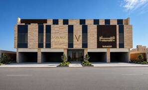 A modern building with 'VOYAGE EXPO SUITES' signage, featuring a prominent 'V' logo, and Arabic script on the right side.