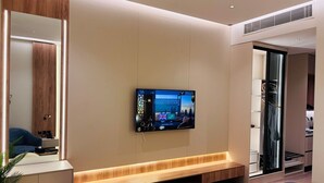 A modern hotel room with a large TV mounted on the wall, a wooden bench, a mirror, and a walk-in closet. - Voyage Expo Suites (Riyadh)