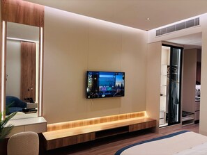 Une chambre d’hôtel moderne dotée d’un grand téléviseur fixé au mur, d’un banc en bois, d’un miroir et d’un dressing.