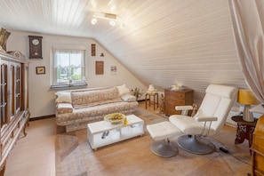 A cozy attic bedroom with a sofa, a white chair, a coffee table, and a dresser.