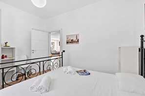 Quarto de hotel com cama, escrivaninha, cadeira, radiador, quadro na parede e porta que dá para outro cômodo.