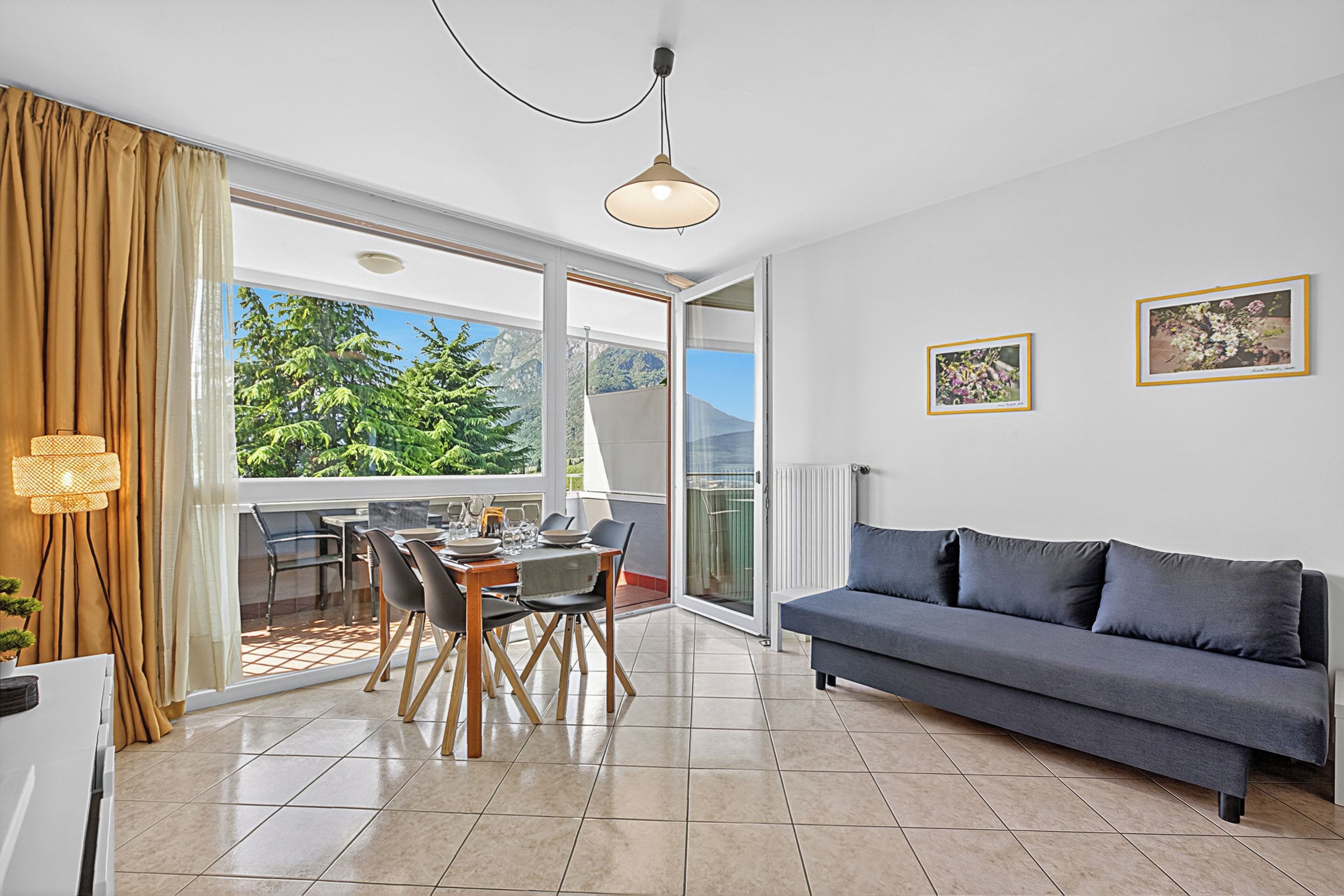 A modern living room with a sofa, dining table, chairs, and a large window with a view of greenery.