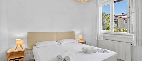 A neatly made bed with white linens, two bedside lamps, a wicker lamp shade, and a window with curtains.