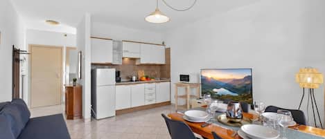 A modern kitchen with white cabinets, a dining area with a wooden table set for two, and a painting of a landscape on the wall.