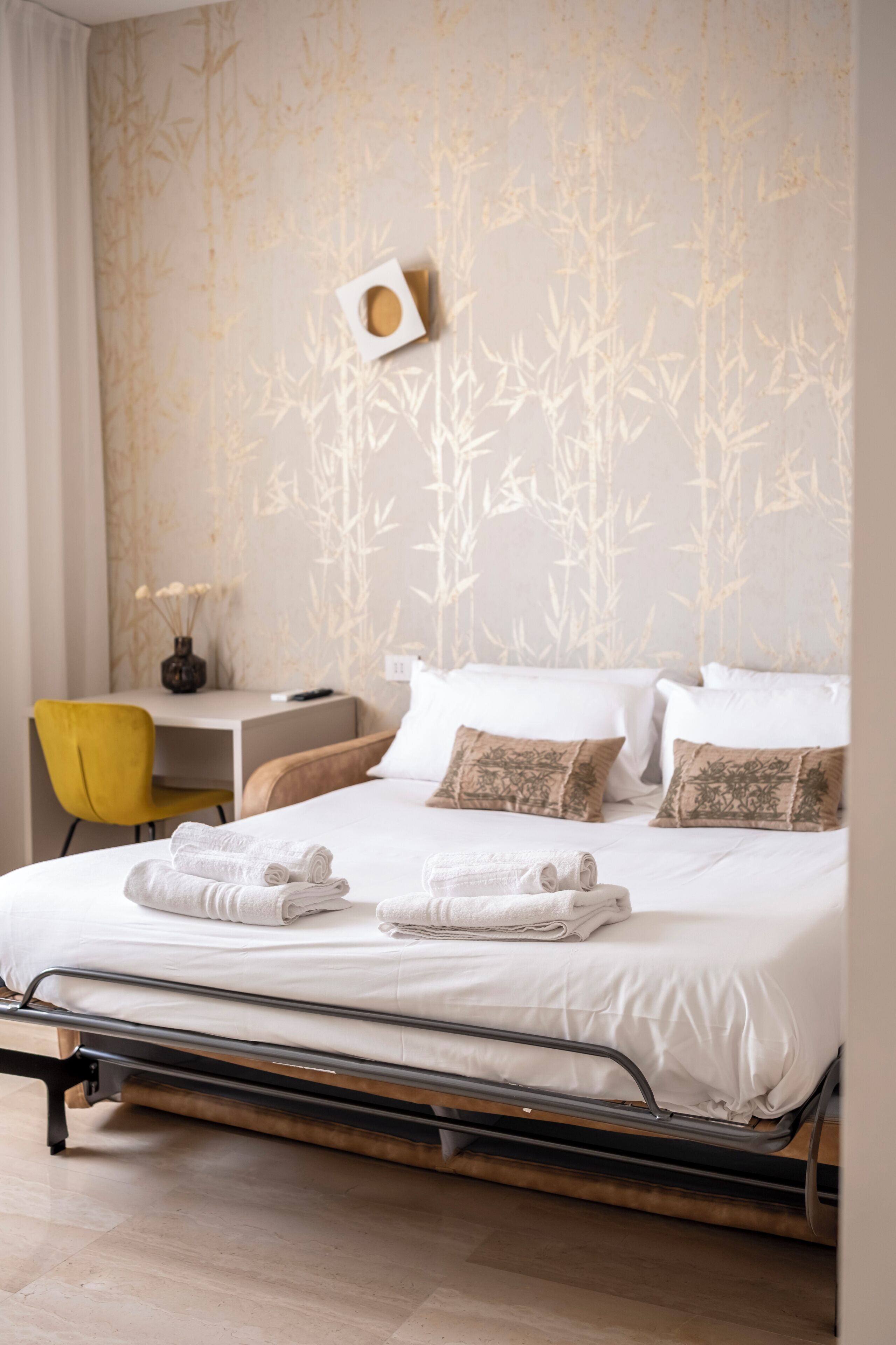 A neatly made bed with white linens, a yellow chair, a small table with a vase, and a wall with a bamboo pattern.