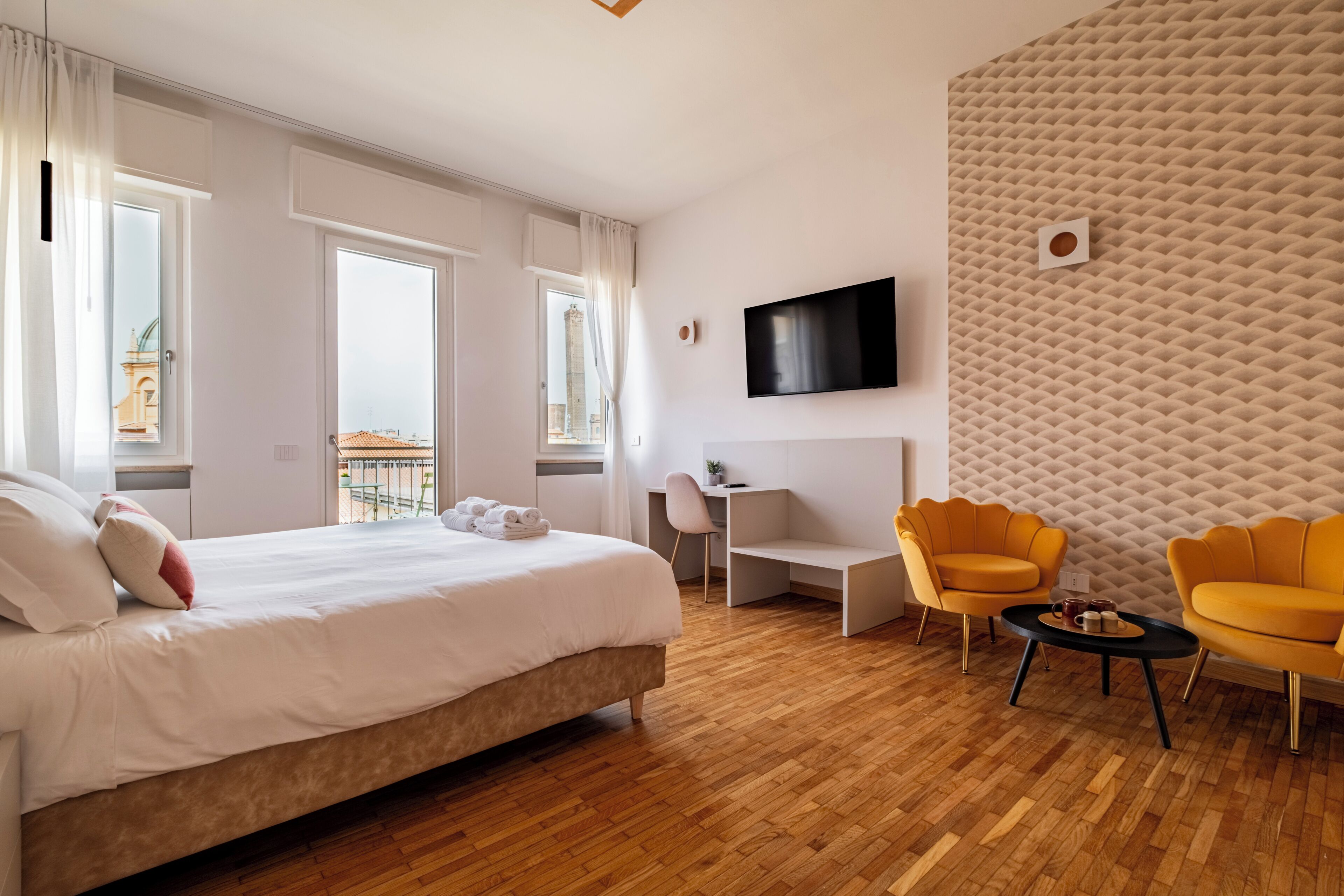 A hotel room with a bed, a desk, two chairs, a TV, and a view of a building outside.