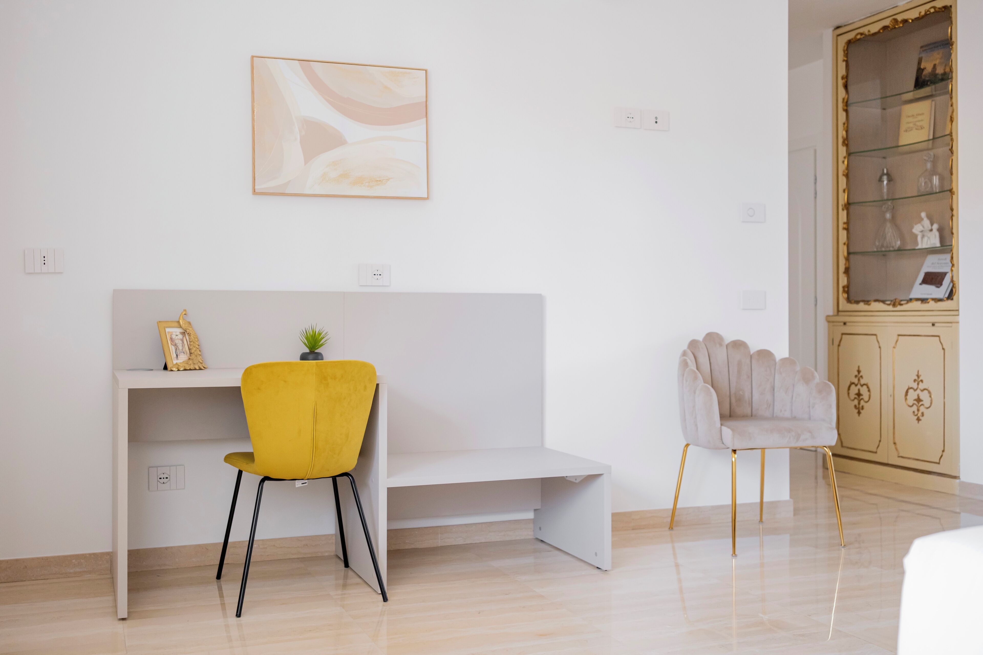 A modern room with a white desk, a yellow chair, a grey armchair, and a cabinet with glass doors.