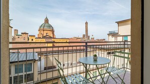 Un balcón con mesa y sillas, una planta en maceta y vistas a un paisaje urbano que incluye una iglesia y una torre.