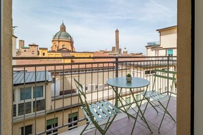 Un balcone con tavolo e sedie, un vaso di fiori e vista su una città con una chiesa e una torre.