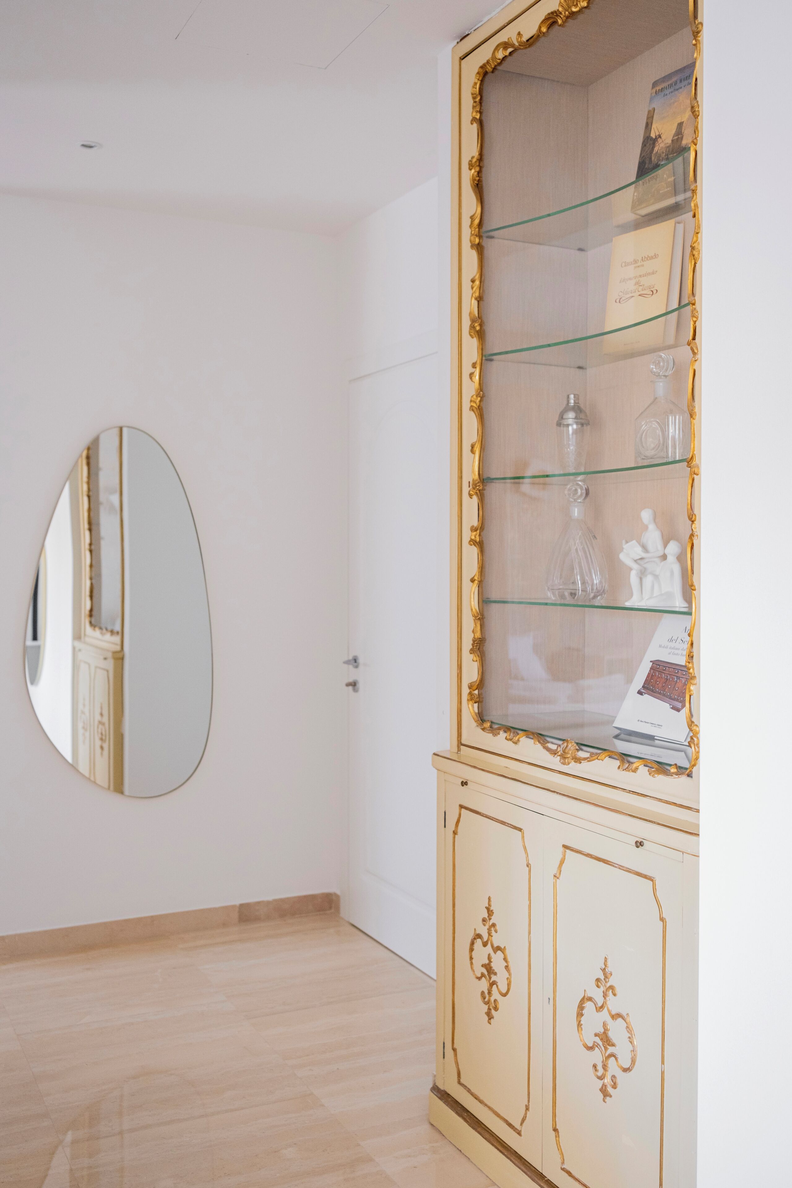 Una habitación con un espejo grande en la pared, un aparador de vidrio con objetos decorativos y suelo de madera.