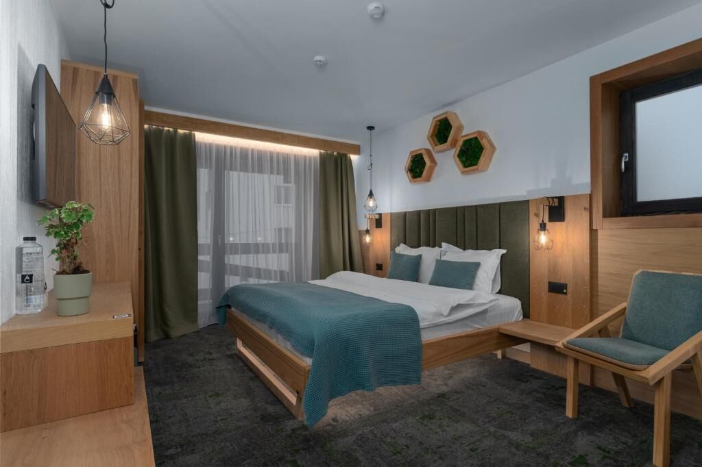 Habitación de hotel moderna con cama, zona de estar, televisor, planta y ventana con cortinas.