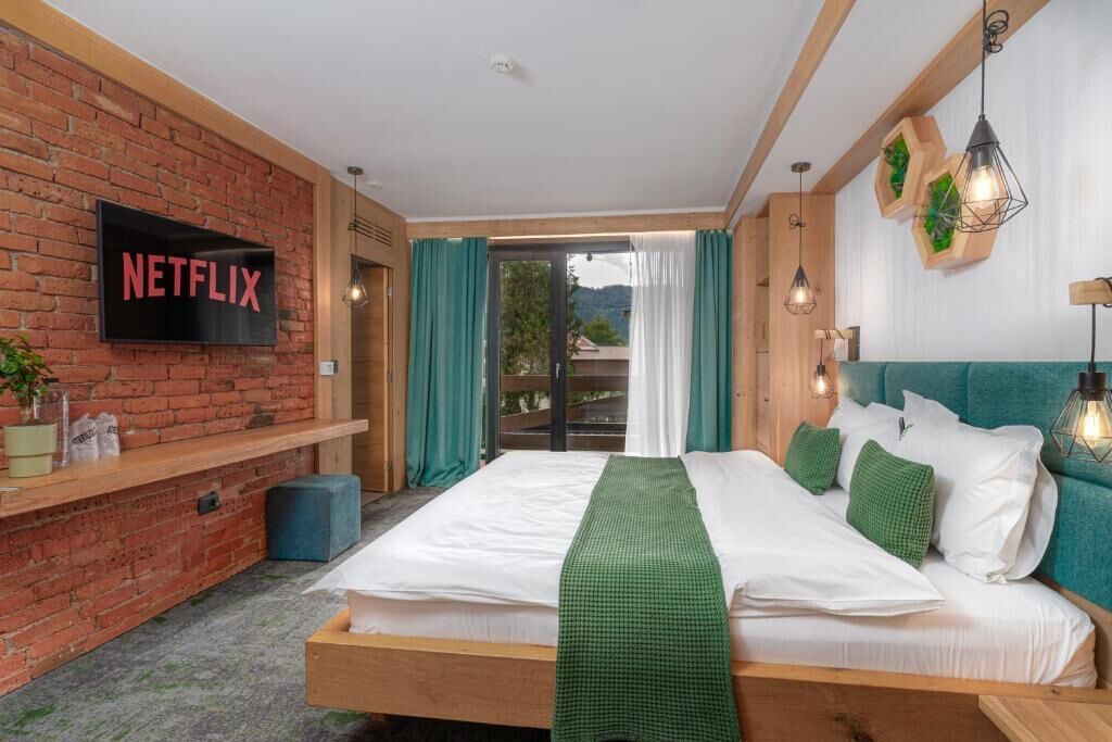 Et moderne hotelværelse med en murstensvæg, et Netflix-skilt, en seng med grønt sengetøj og udsigt gennem en glasdør.