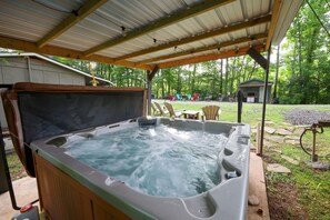 A hot tub under a covered patio with a wooden deck, surrounded by a grassy yard and trees. - BabyBears Peaceful cabin & hot tub (Whittier)