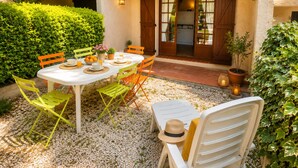 Um pátio externo com uma mesa posta para refeições, rodeado de cadeiras, e uma porta que dá para um edifício.