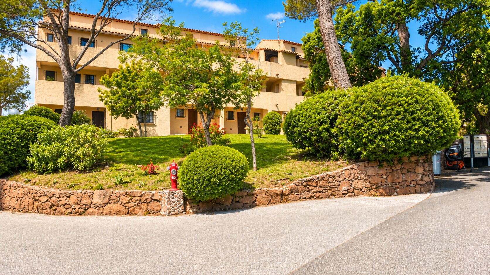 Un edificio a più piani con un muro di sostegno in pietra, un idrante rosso e una vegetazione curata.