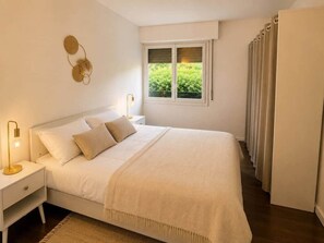 Ein Schlafzimmer mit Bett, Nachttischen, einer Lampe, einem Fenster mit Jalousien und einer Tür mit Vorhängen.