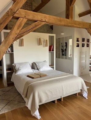 A bedroom with a large bed, wooden beams, a bookshelf, and a mirror. - La Maison de la Tour (Léré)