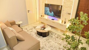 A modern living room with a beige sofa, a round coffee table, a flat-screen TV, and a potted plant. - Eleven Apartments (Galadimawa)