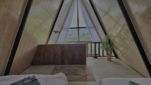 A triangular tent-style room with two beds, a wooden bench, a view of the landscape, and a potted plant.