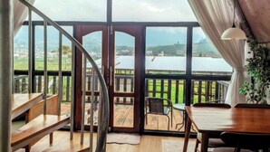 A room with a wooden staircase, a dining area with a table and chairs, and a large window offering a view of a body of water and greenery.