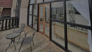 A modern building with a large glass facade, wooden deck with metal chairs, and a visible door.