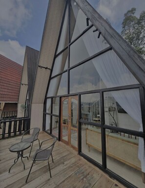 Un edificio moderno con una gran fachada de vidrio, terraza de madera con sillas metálicas y una puerta visible.