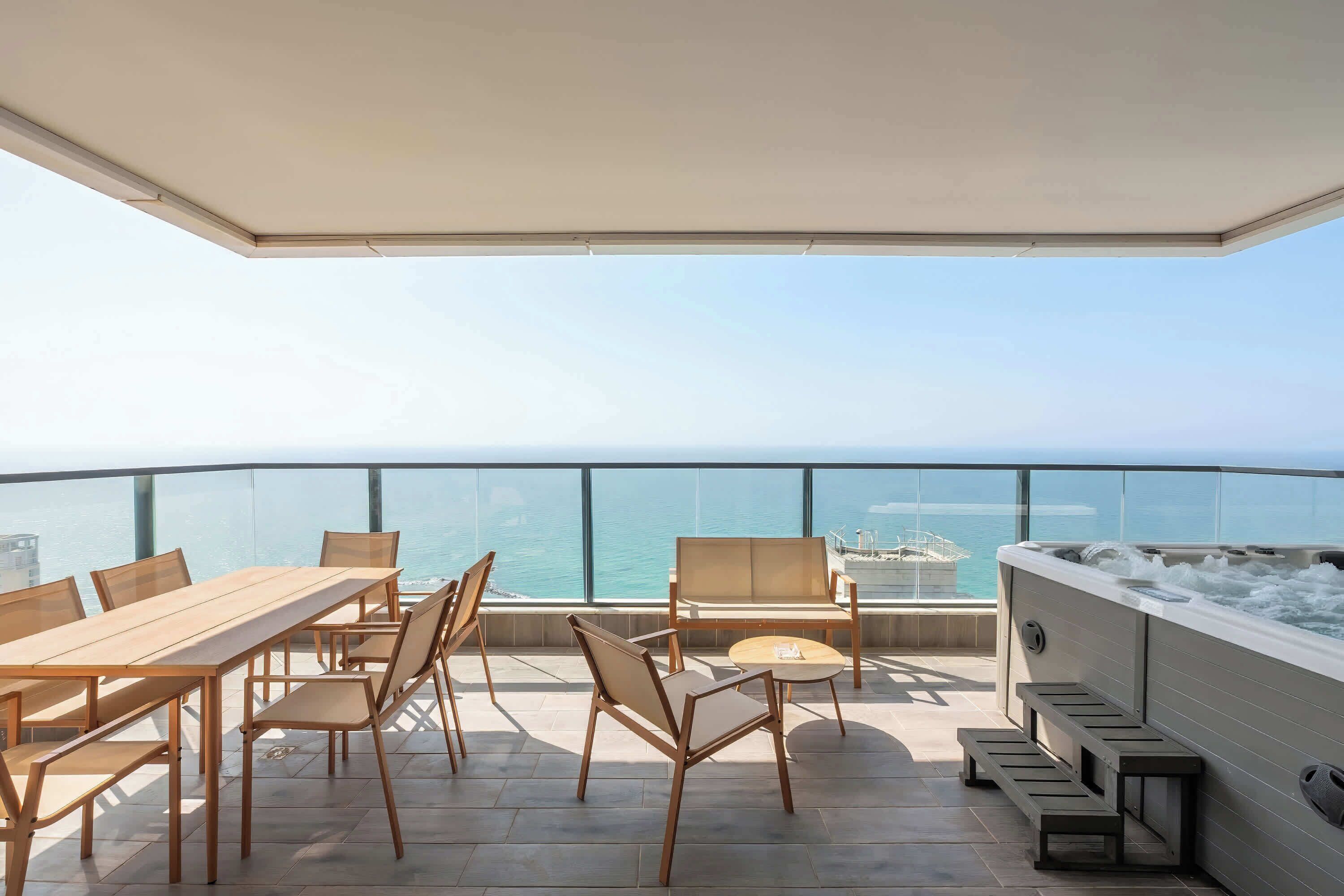 Eine Dachterrasse mit Holztisch und -stühlen, einem Whirlpool und einem Glasgeländer mit Blick aufs Meer.