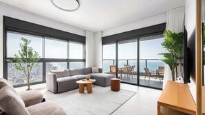 Een moderne woonkamer met een grijze hoekbank, een ronde salontafel en een groot raam met uitzicht op de stad.