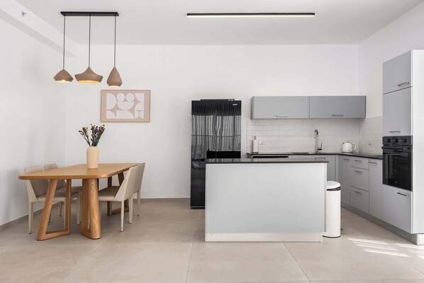 A modern kitchen with a central island, pendant lighting, and a dining area with a wooden table and chairs. - Luxury 5BR PH Balcony Jacuzzi w Mamad (Bat Yam)