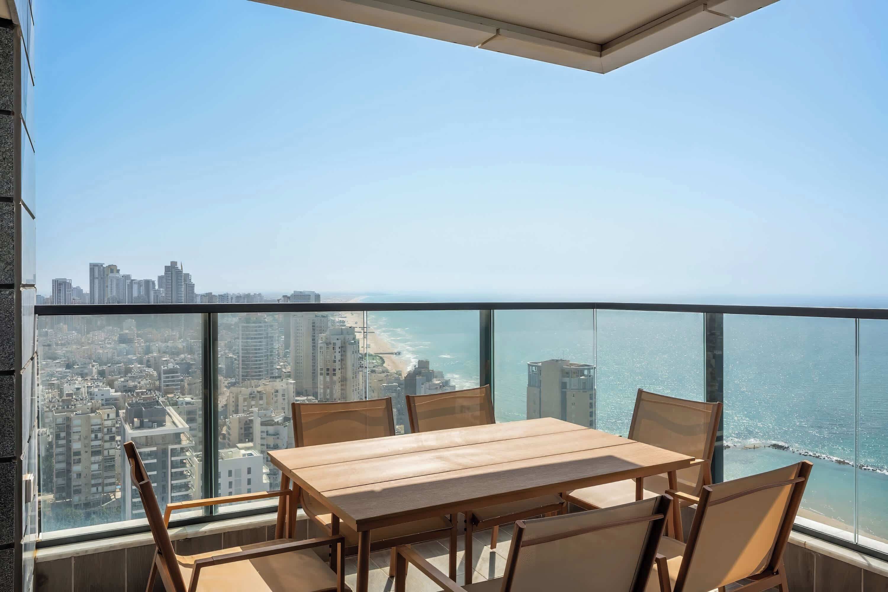 A balcony with a wooden table and chairs, overlooking a cityscape and the ocean.